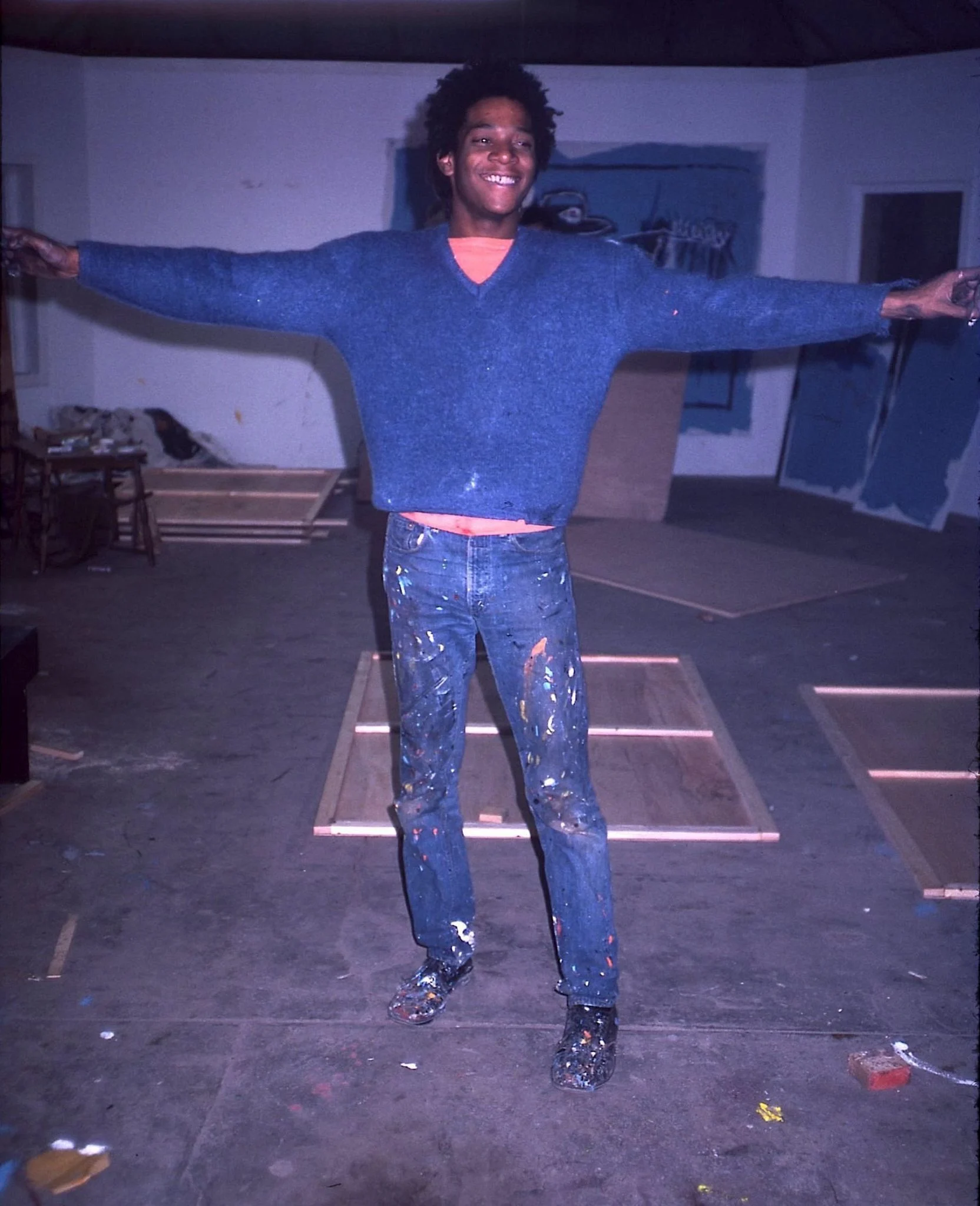 Jean-Michel Basquiat in his studio, around 1981, paint-spattered, arms outstretched