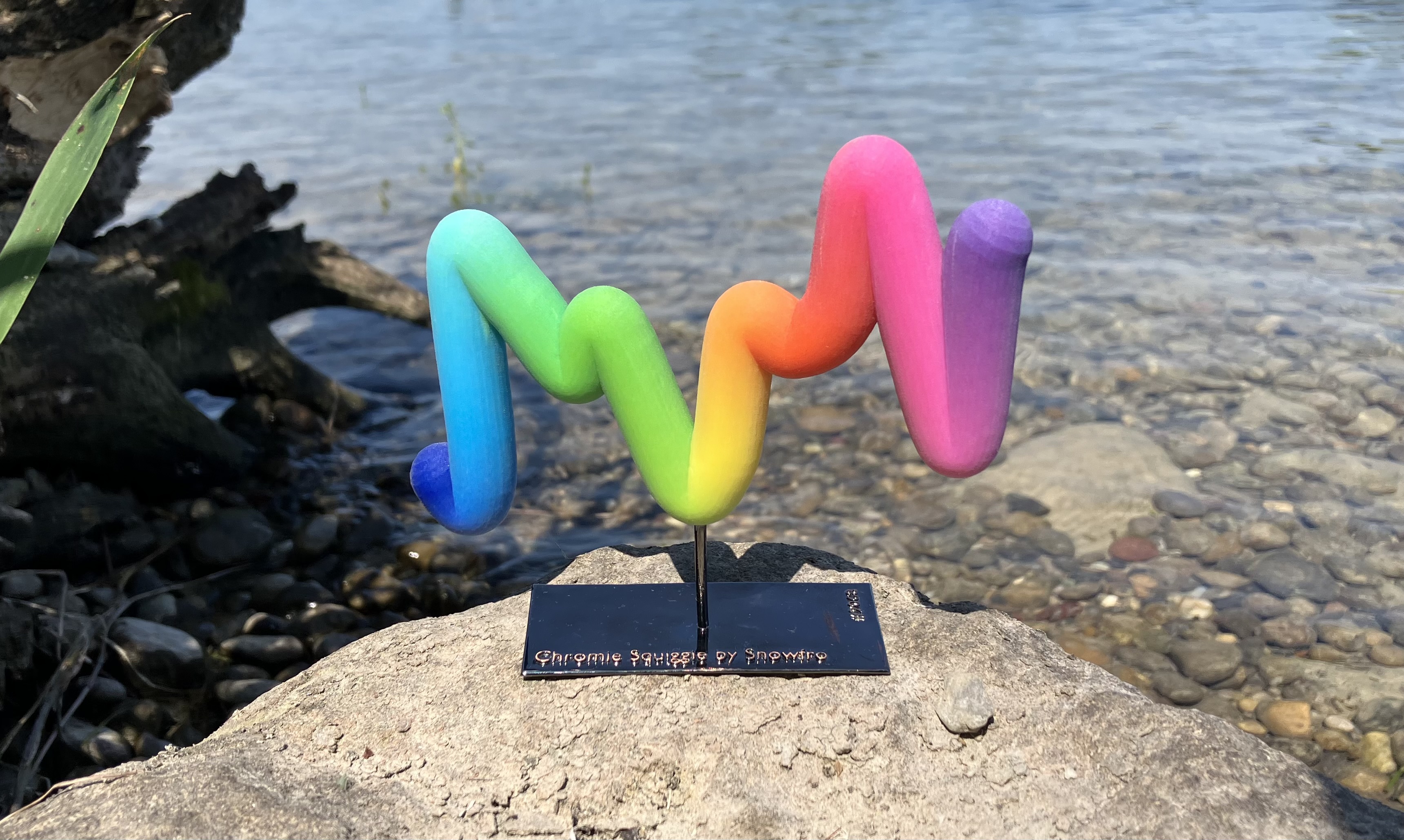 Charley’s rainbow Chromie Squiggle sculpture on a black stand, photographed on a rock beside clear water